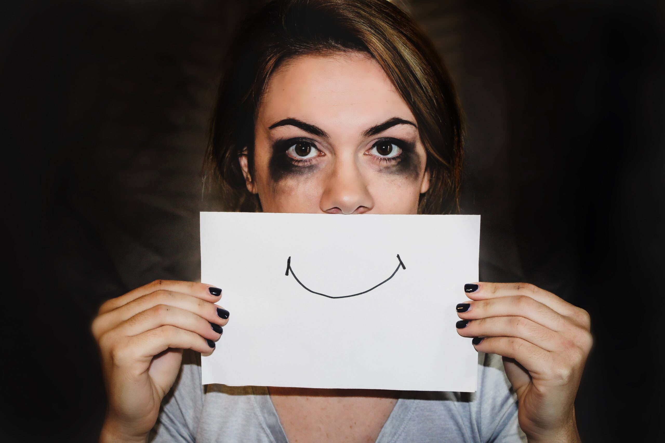 Image of women tired with mascara smudged around her eye, looking stressed, holding a piece of paper with a smimley face on it in front of her mouth.